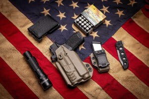 Golden Saber Bonded box and cartridges laying on an American flas with a flashligh, handgun in a holster, handgun magazine, and knife